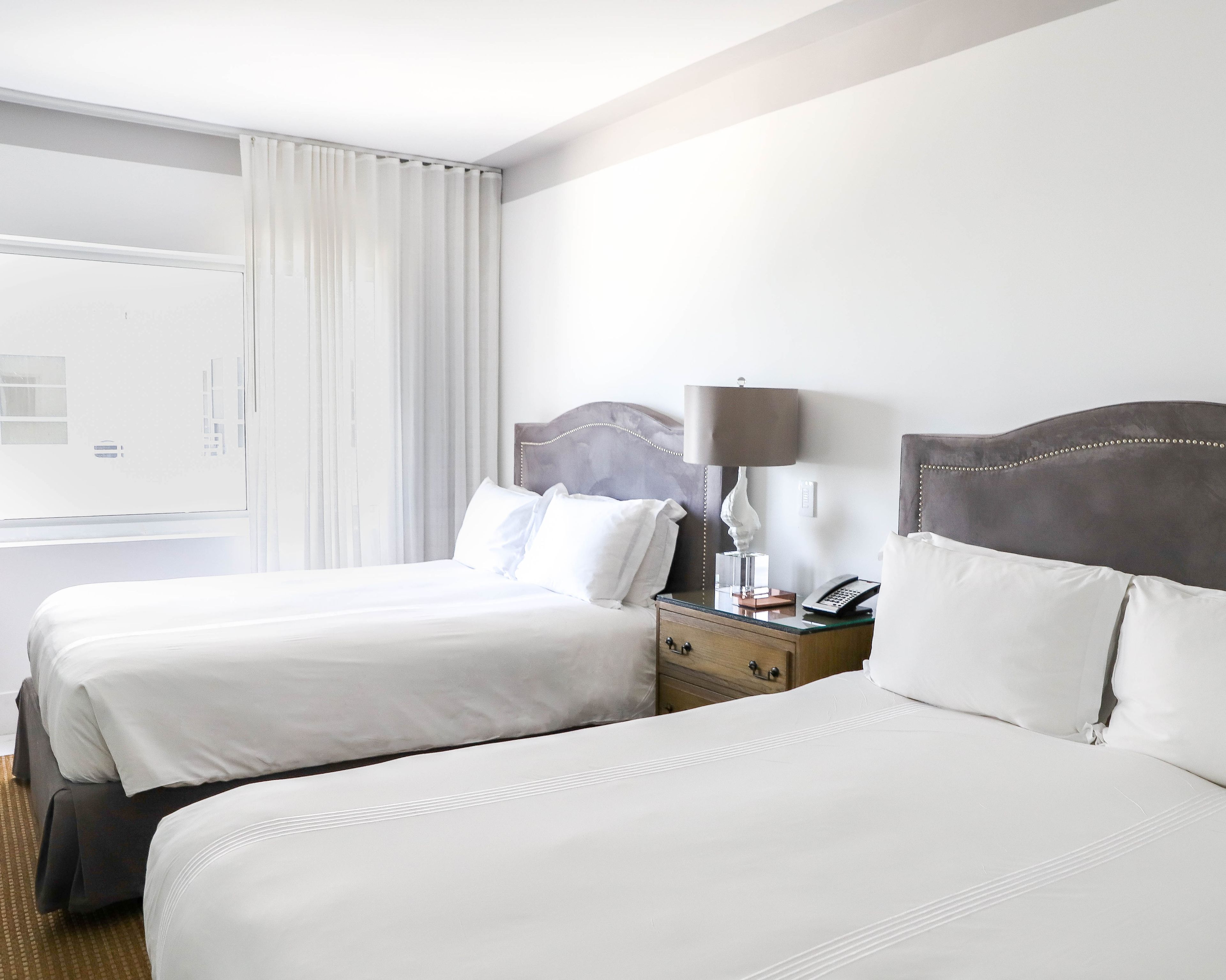 A hotel room with two beds, a nightstand with a lamp, and a window with curtains.