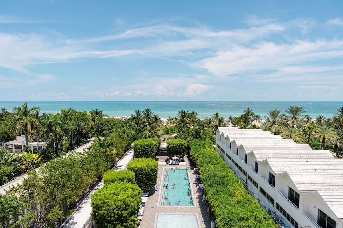 A resort with a swimming pool surrounded by lush greenery and palm trees, overlooking a beach.