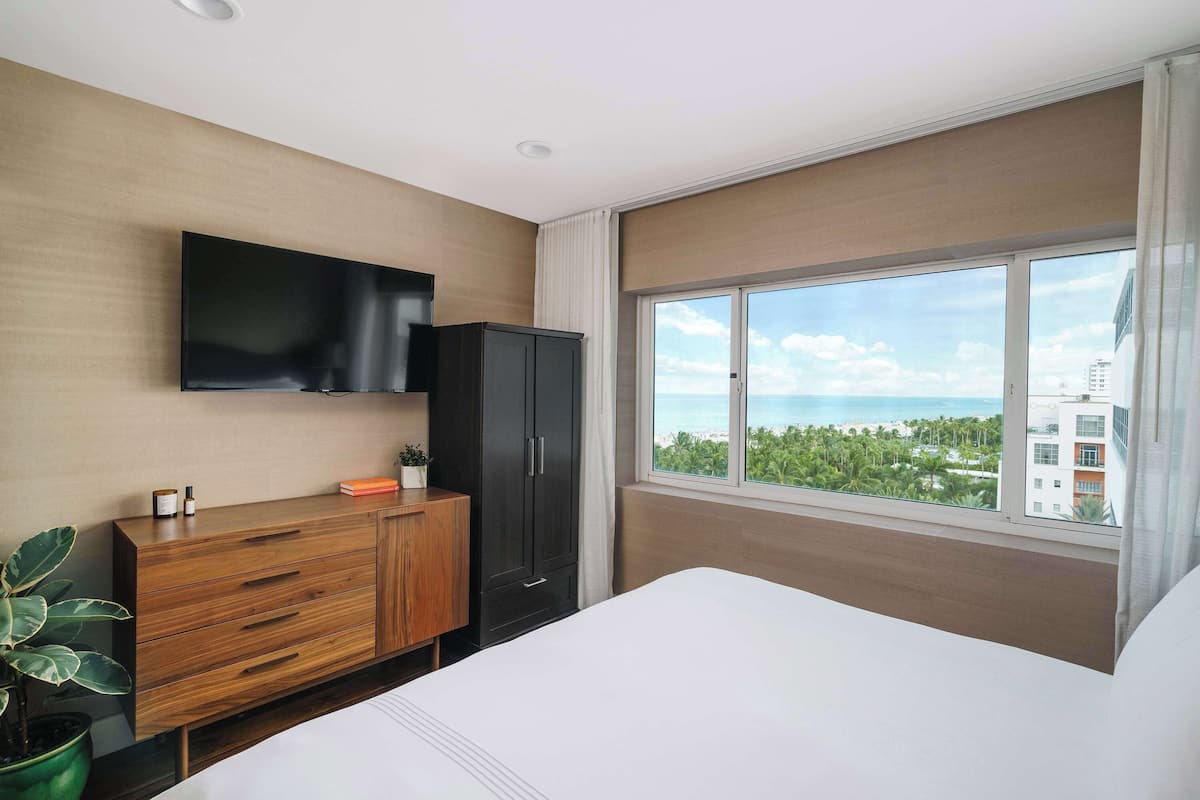 A modern hotel room with a large bed, a wooden dresser, a flat-screen TV mounted on the wall, and a large window offering a view of the ocean and buildings.