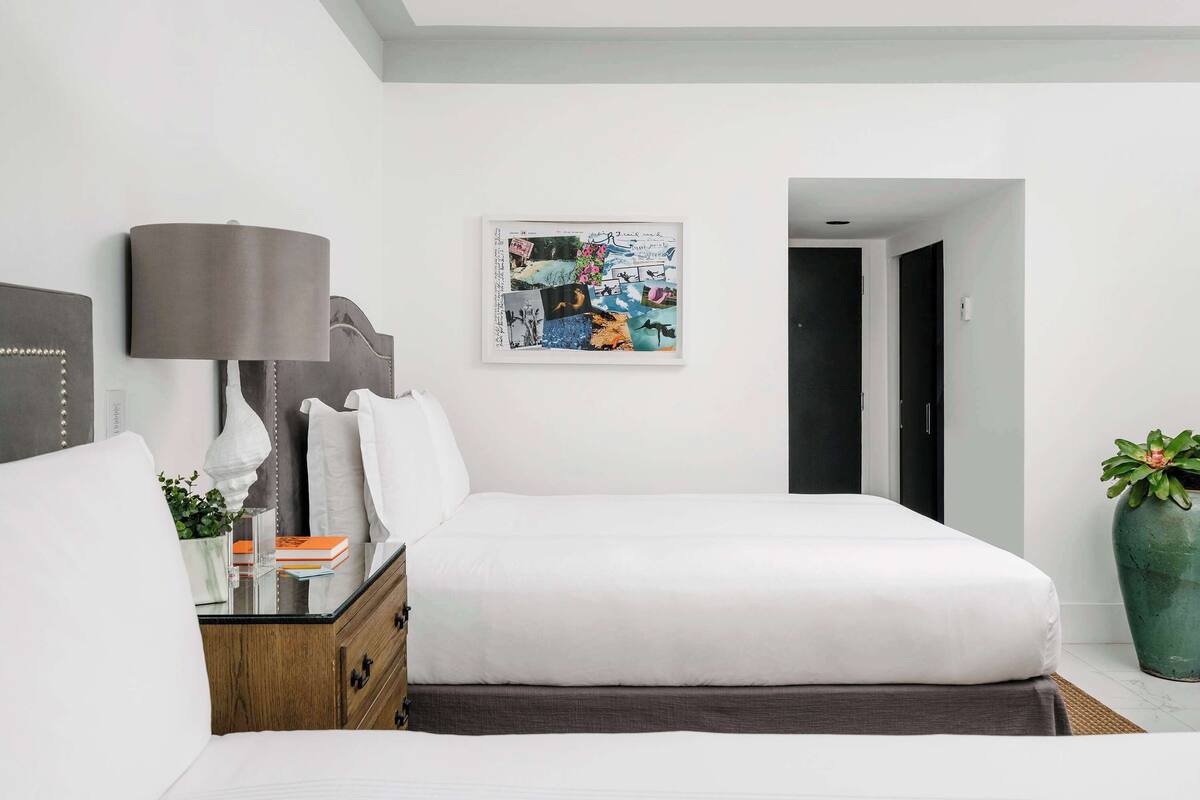 A modern hotel room with a large bed, a bedside table with a lamp, a wall art piece, and a potted plant.