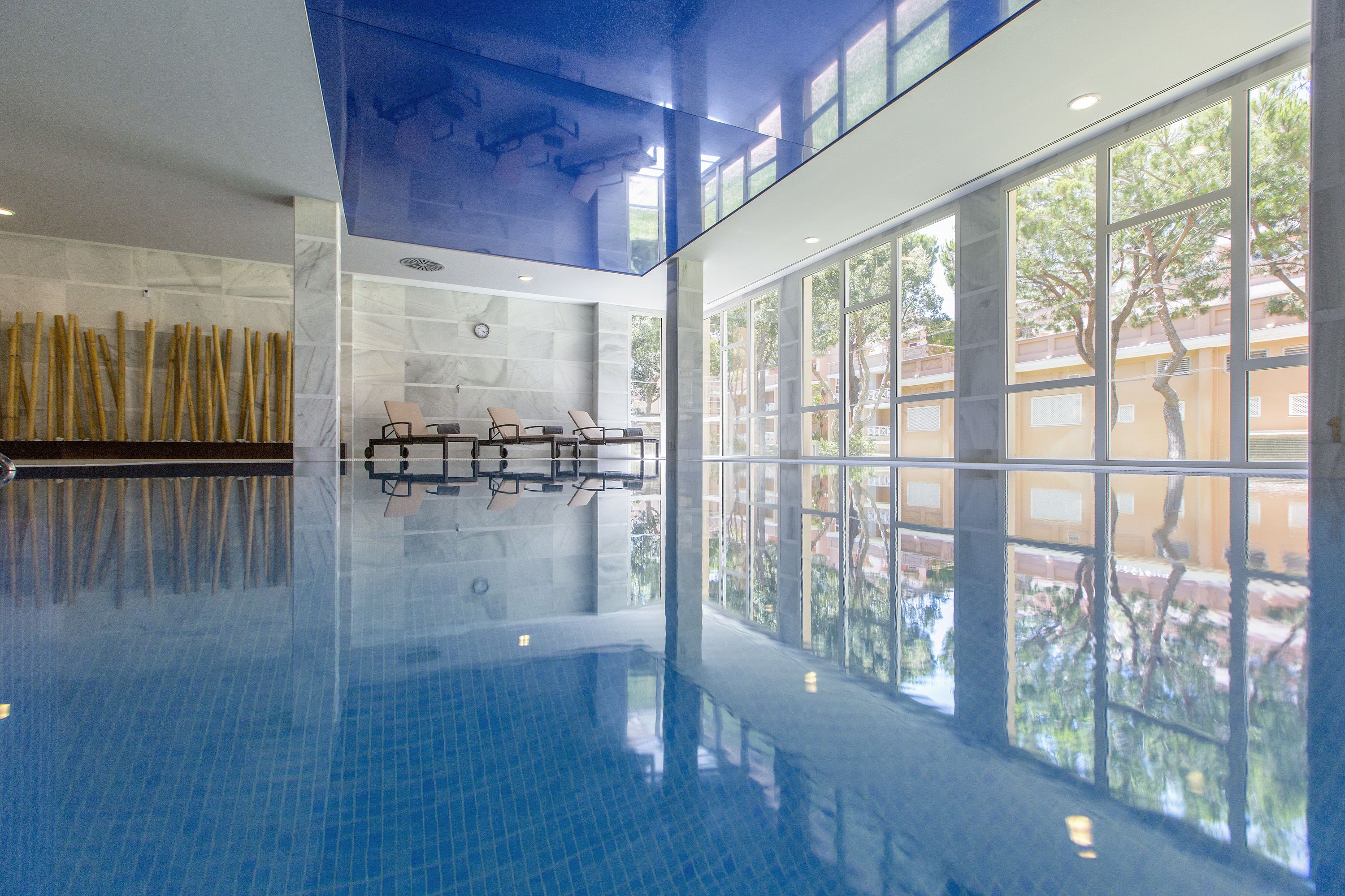 A large indoor swimming pool with clear glass walls, a tiled floor, and a seating area with chairs.