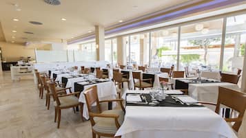 Un comedor amplio con varias mesas dispuestas para comer, ventanas grandes que proporcionan luz natural y una pizarra al fondo.