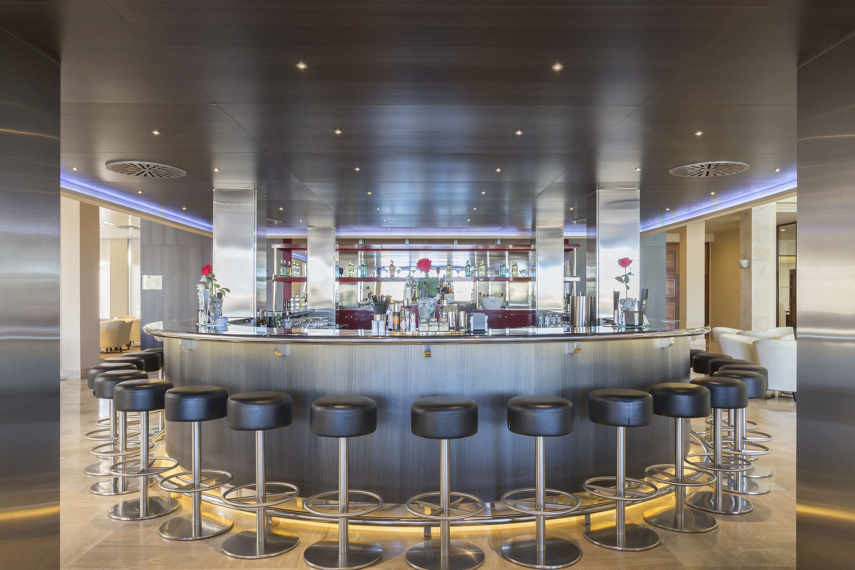 A modern bar area with a central bar, multiple stools, and a well-lit ceiling.