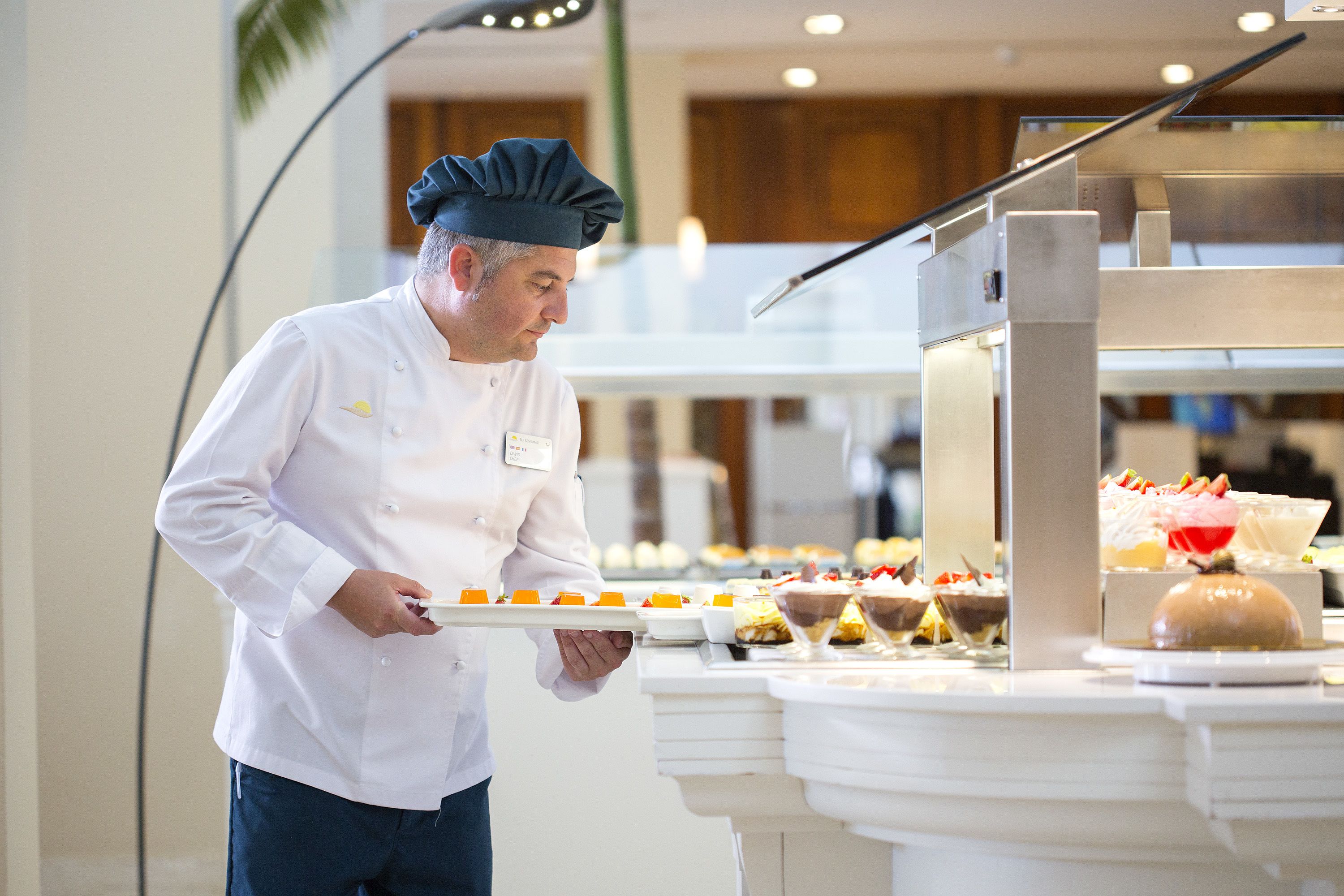 A chef in a white uniform holding a tray of desserts in a buffet setting.
