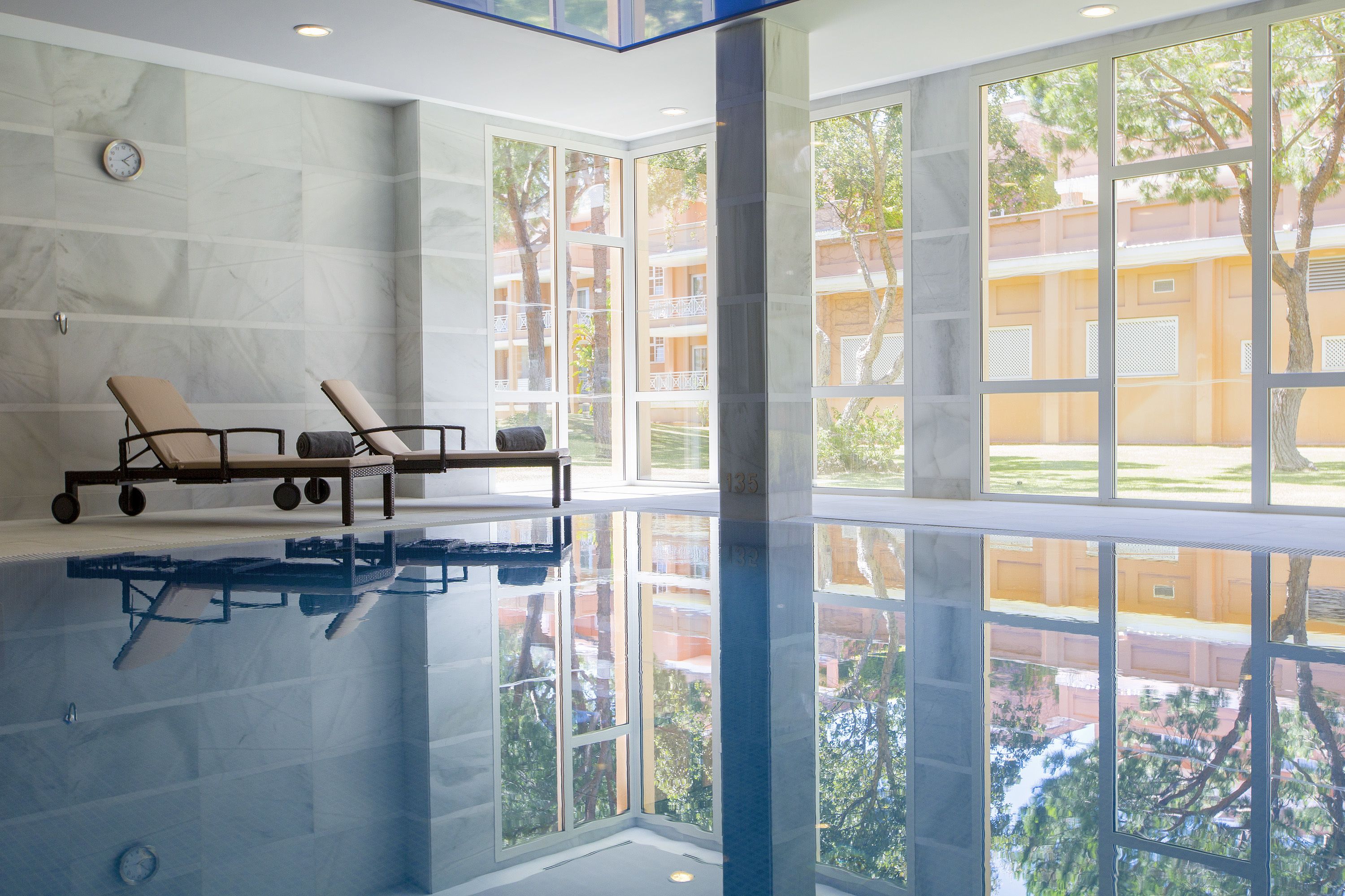 A spacious indoor pool area with clear glass walls, a lounge with chairs, and a visible clock on the wall.
