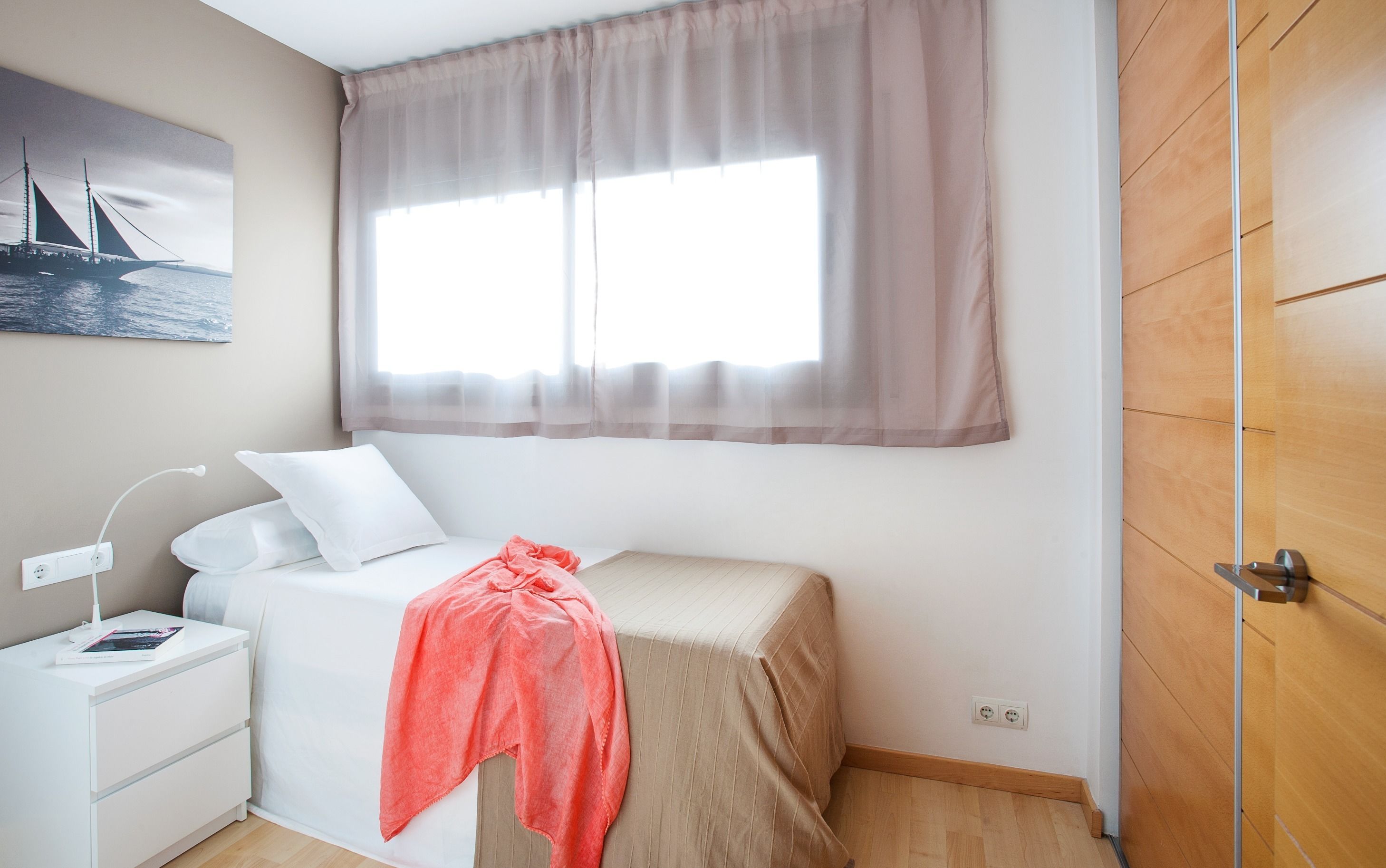 A small, neatly arranged bedroom with a single bed, a bedside table with a lamp, a power outlet, and a window with sheer curtains.