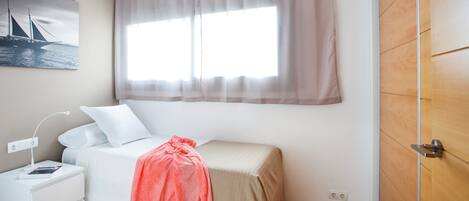 A small, neatly arranged bedroom with a single bed, a bedside table with a lamp, a power outlet, and a window with sheer curtains.