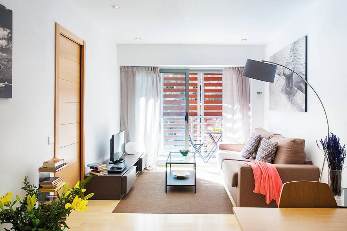 Una sala de estar moderna con un sofá, una mesa de centro y un televisor. Hay una estantería con libros y una planta.