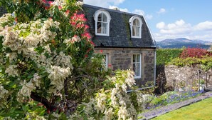 A stone house with a chimney, white-framed windows, and a garden with various plants and flowers. - Leven House (Crieff)