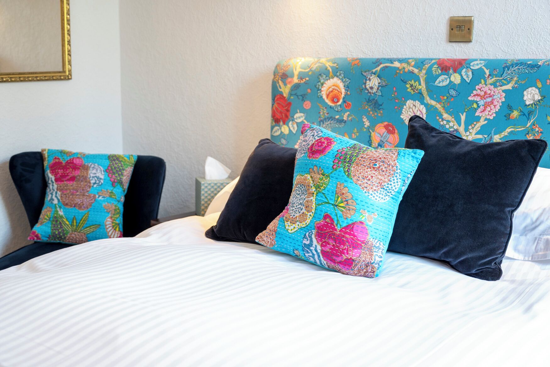 A bed with floral-patterned pillows, a bedside table with a tissue box, and a wall-mounted light switch.