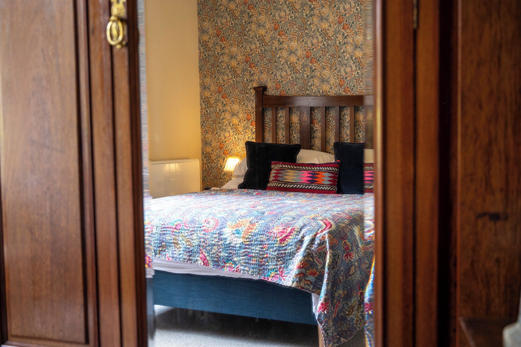 A bedroom with a patterned wallpaper, a wooden headboard, and a bed with a colorful quilt.