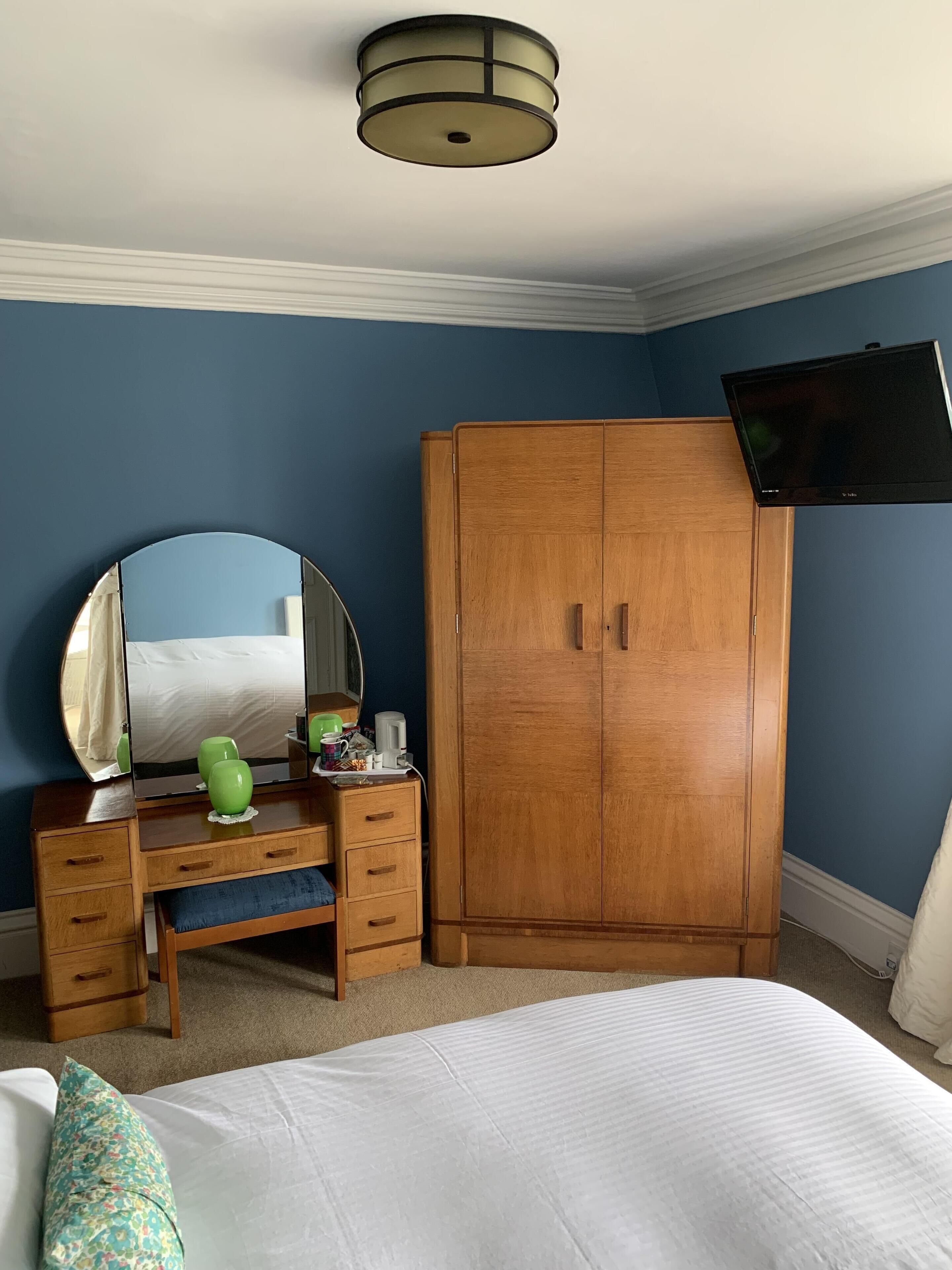 Une chambre à coucher avec une commode en bois, un miroir, un téléviseur fixé au mur et un lit recouvert d’une couette blanche.
