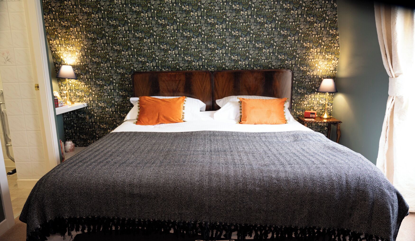 A bed with a dark patterned bedspread and two orange pillows, a wooden headboard, and a wall with a floral wallpaper pattern.