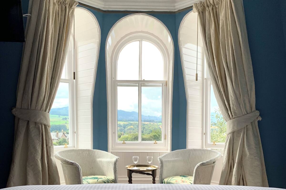 Un dormitorio con una cama grande, dos sillas blancas, una mesita con vasos y una ventana con cortinas.