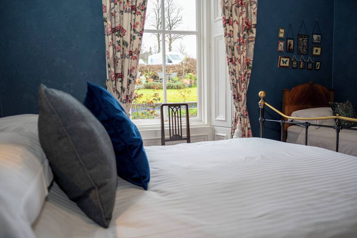 Un dormitorio con cama, una silla, un ventanal con cortinas y una pared decorada con marcos.