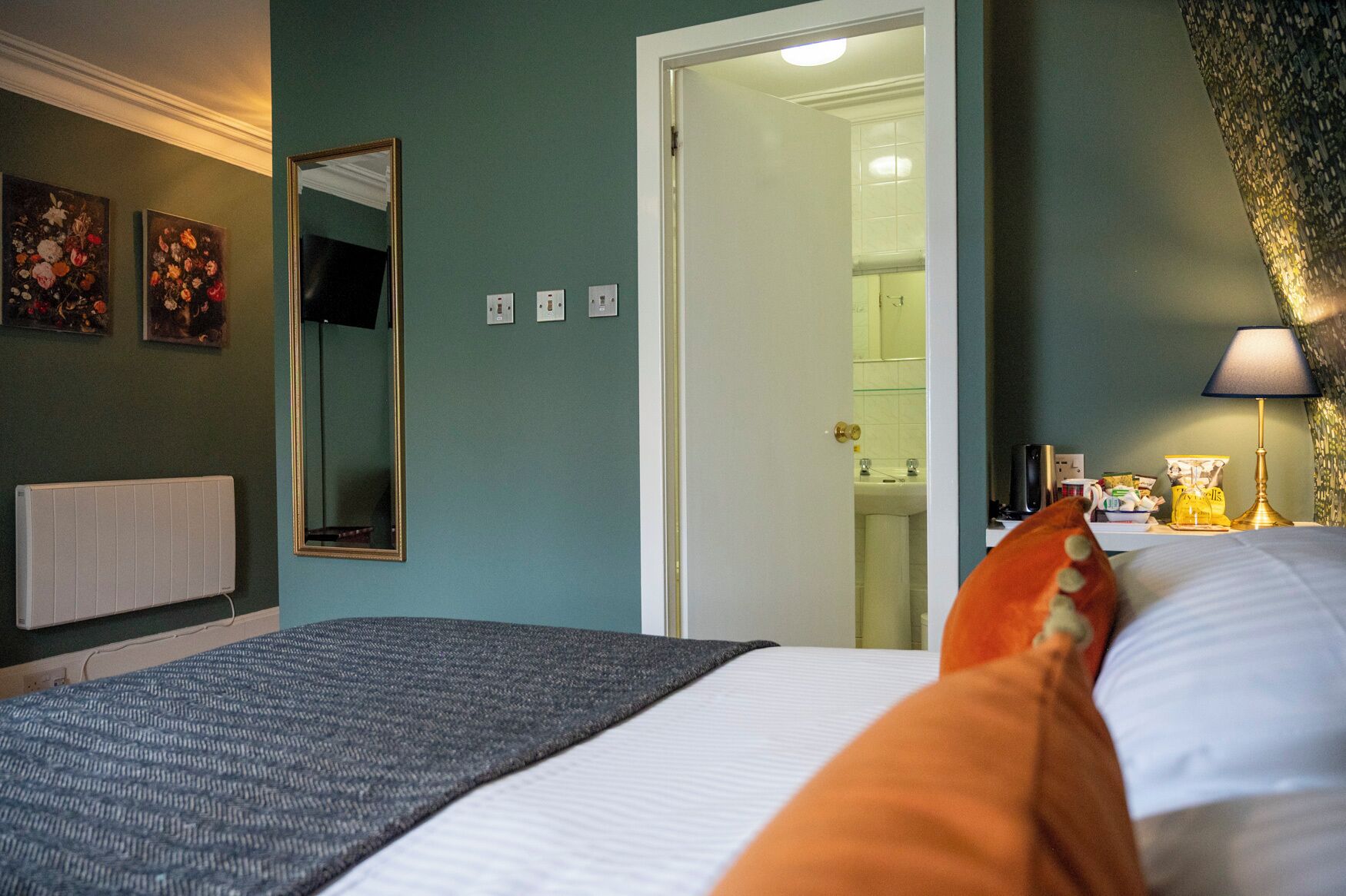 A bedroom with a bed, a nightstand, a lamp, a mirror, and a door leading to a bathroom.