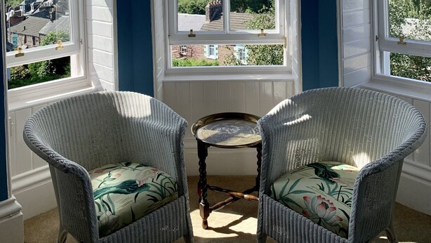 A room with a bay window overlooking a scenic landscape, two wicker chairs with floral cushions, and a small round table. - Leven House (Crieff)
