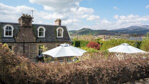 A stone building with arched windows, a chimney, and a view of rolling hills and a lake. - Leven House (Crieff)