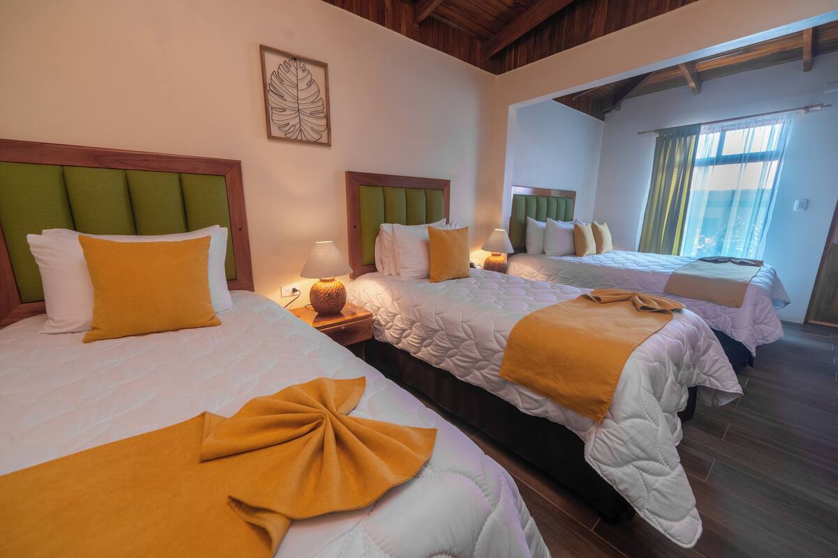 A hotel room with three beds, each with a green headboard, white bedding, and a yellow accent pillow. There are bedside lamps and a decorative item on the nightstand.