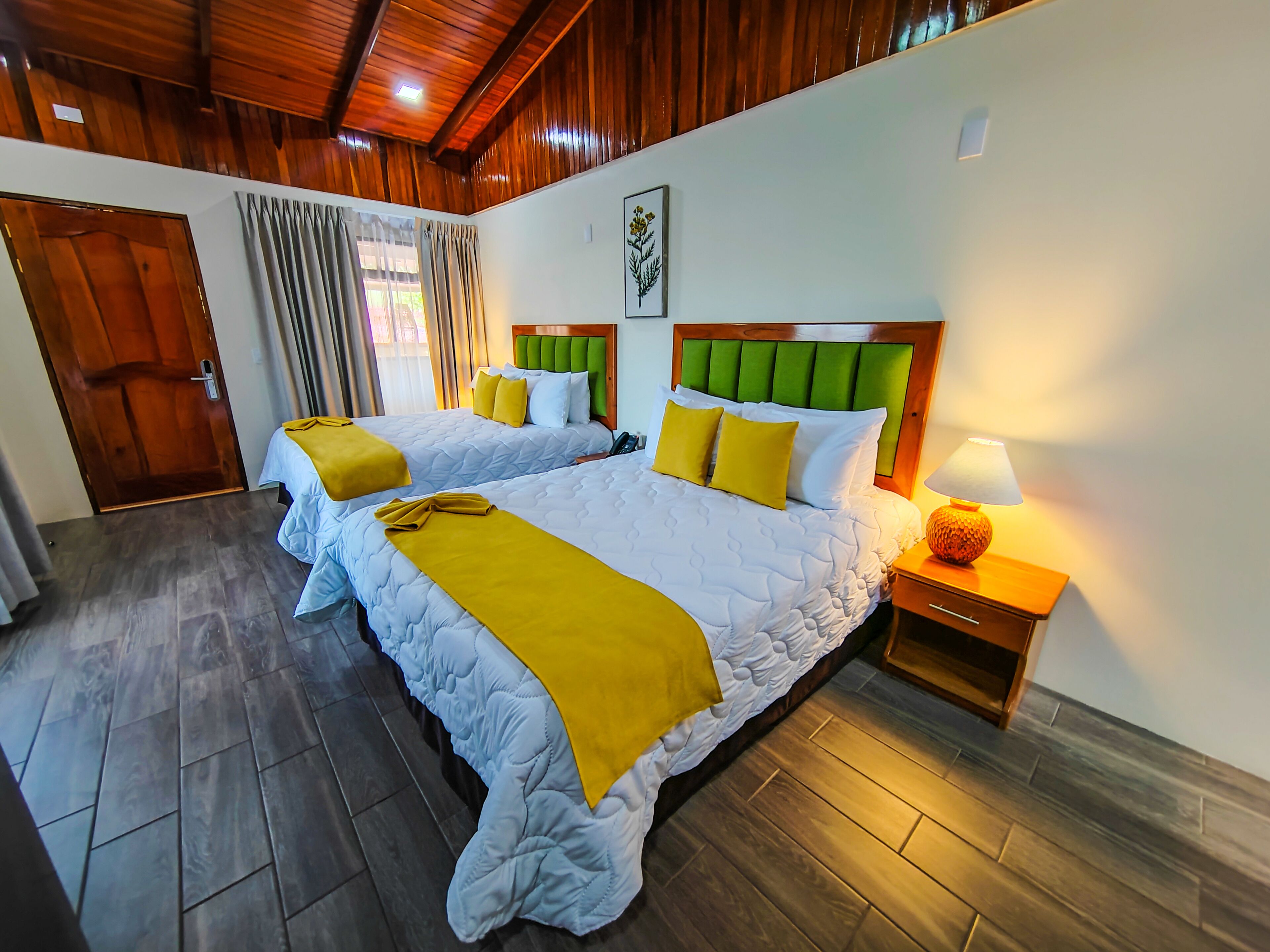 A hotel room with two beds, wooden flooring, a wooden ceiling, and a nightstand with a lamp.