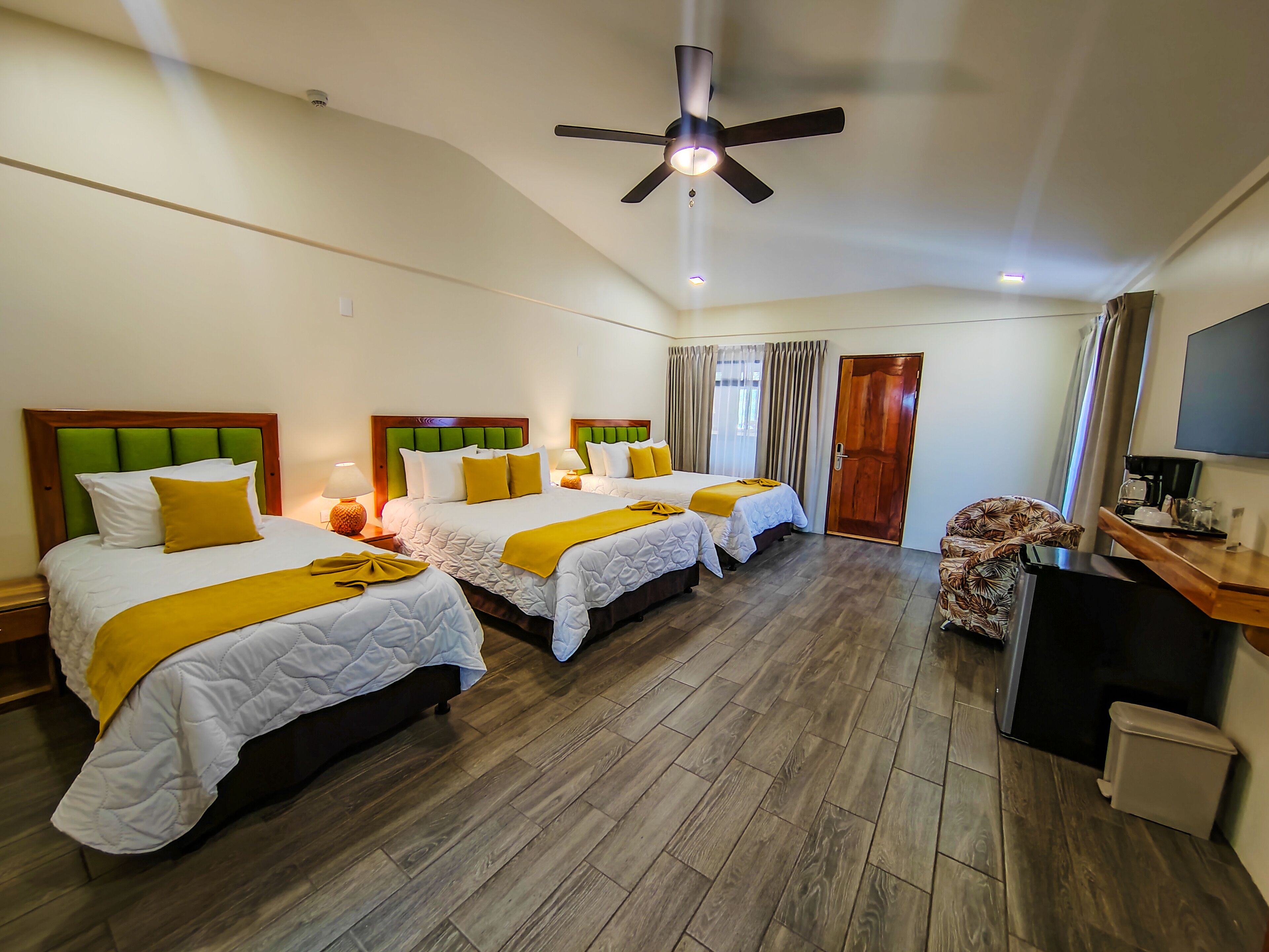 Habitación de hotel con tres camas, ventilador de techo, televisor y cafetera.