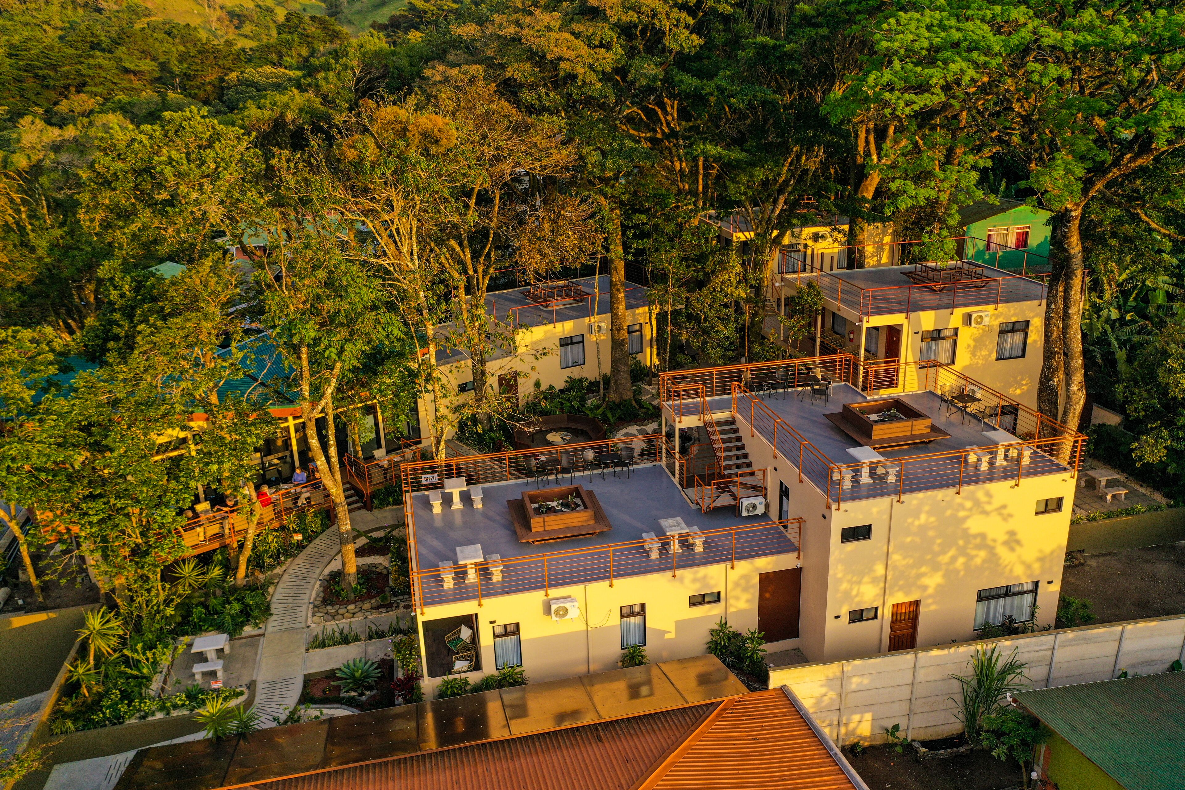Un edificio de varios pisos con una terraza en la azotea, rodeado de un bosque frondoso.