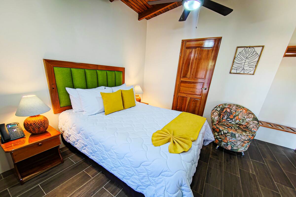 A neatly made bed with a green headboard, a wooden nightstand with a lamp, a floral armchair, and a wooden door.