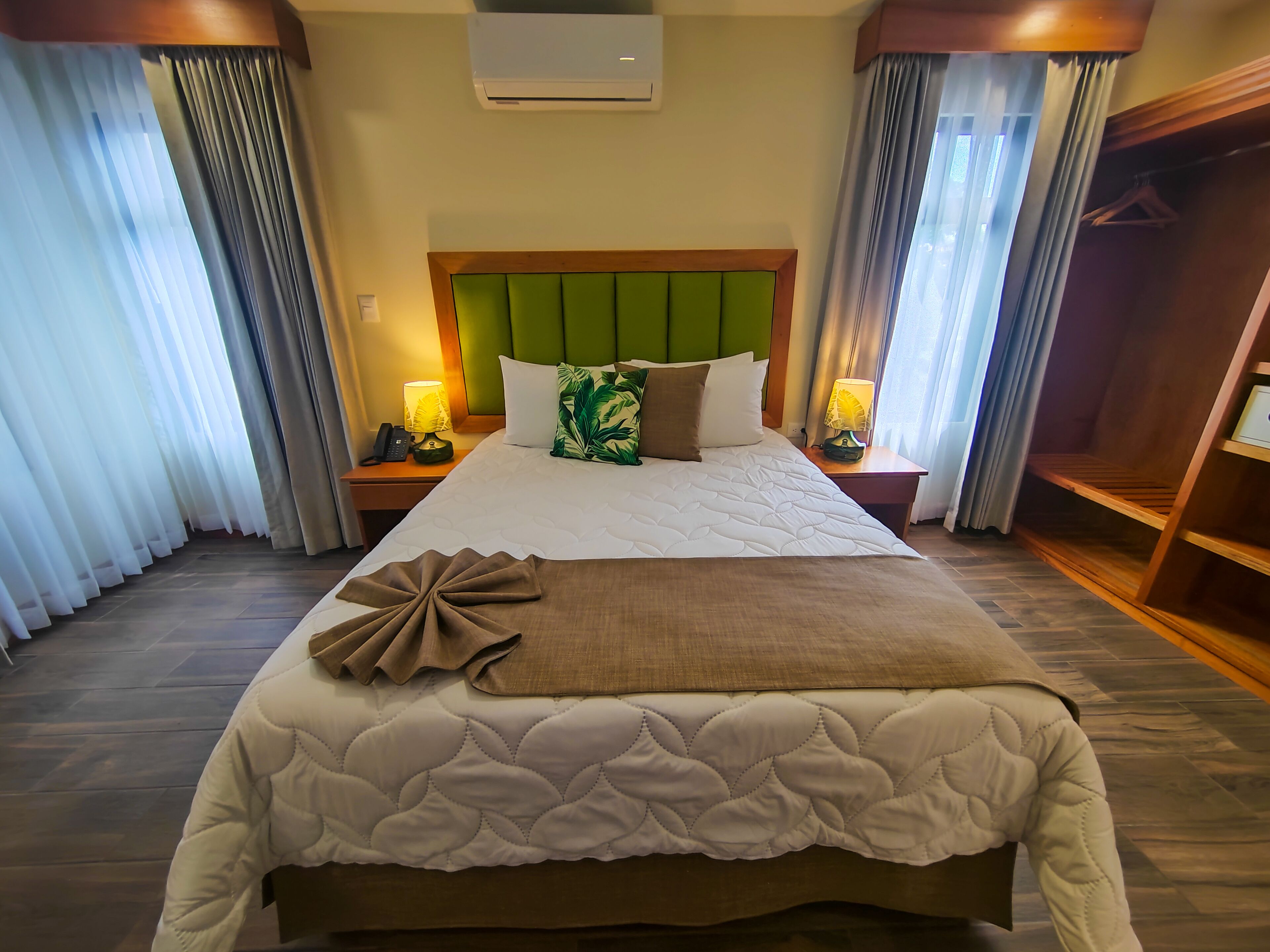 Habitación de hotel con una cama grande, cabecera verde, dos lámparas de noche, una mesita de noche de madera y un armario.