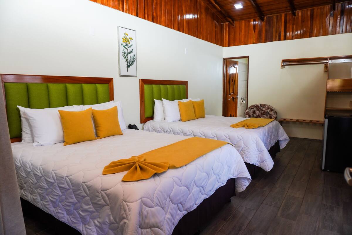 A hotel room with two beds, each with a green headboard and white bedding. There is a wooden ceiling and a door leading to another room.