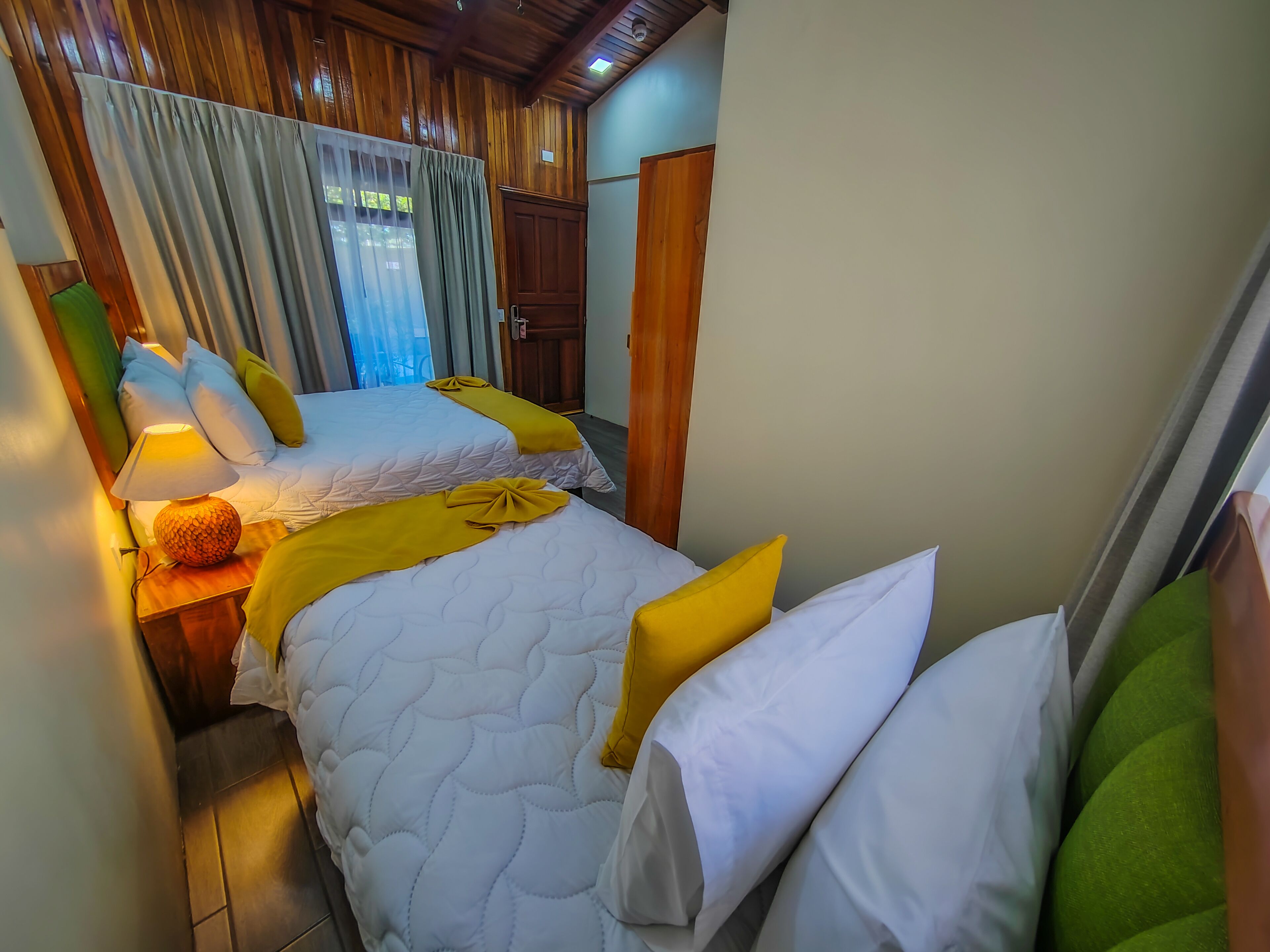 Habitación de hotel con dos camas, paneles de madera, una ventana con cortinas y una mesita de noche con lámpara.
