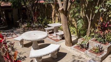 Un patio con una mesa redonda y bancos, rodeado de plantas en macetas y un árbol.