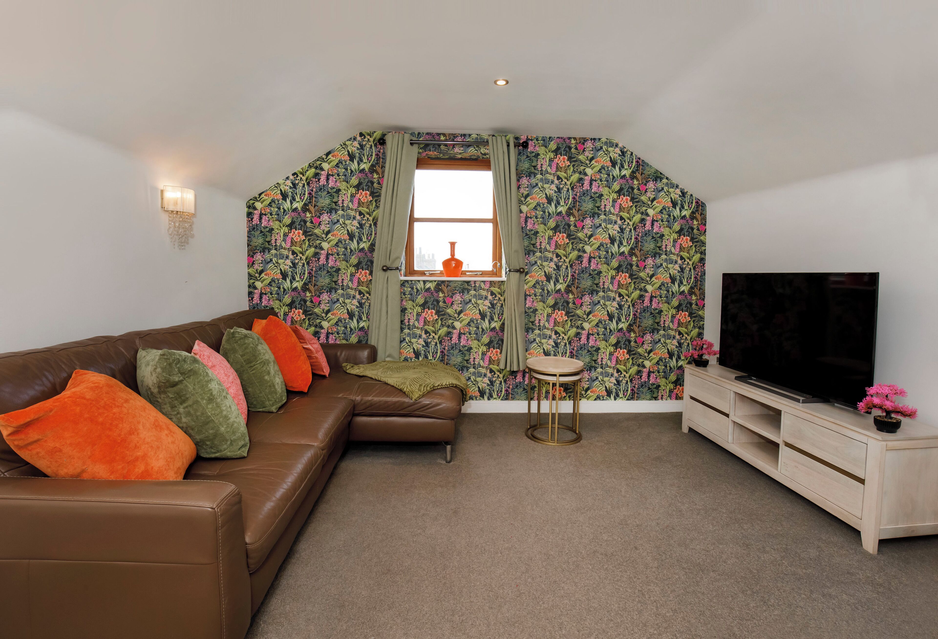 A living room with a brown leather sofa, colorful cushions, a small round table, a TV on a wooden stand, and a window with floral wallpaper.