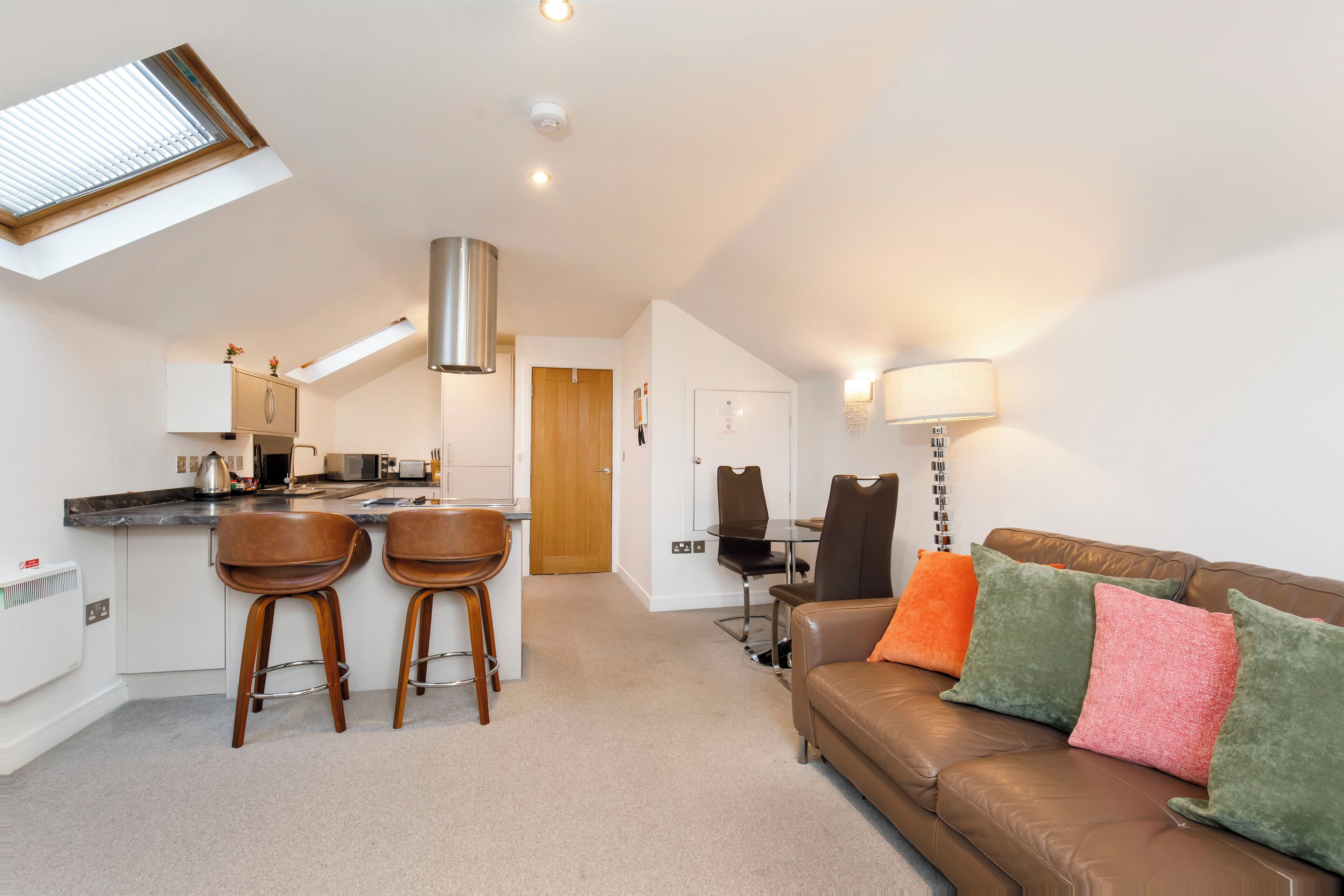 A modern kitchen with a skylight, a dining area with a table and chairs, and a brown leather sofa with colorful cushions.