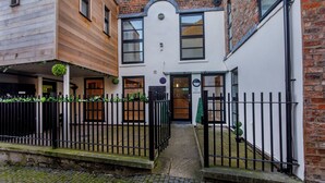 A narrow alleyway with a black metal fence, a white building with a brick facade, and a wooden building on the left.