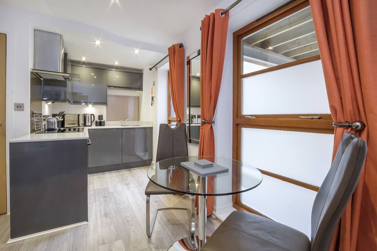 A modern kitchen with a central island, a dining area with a glass table, and orange curtains.