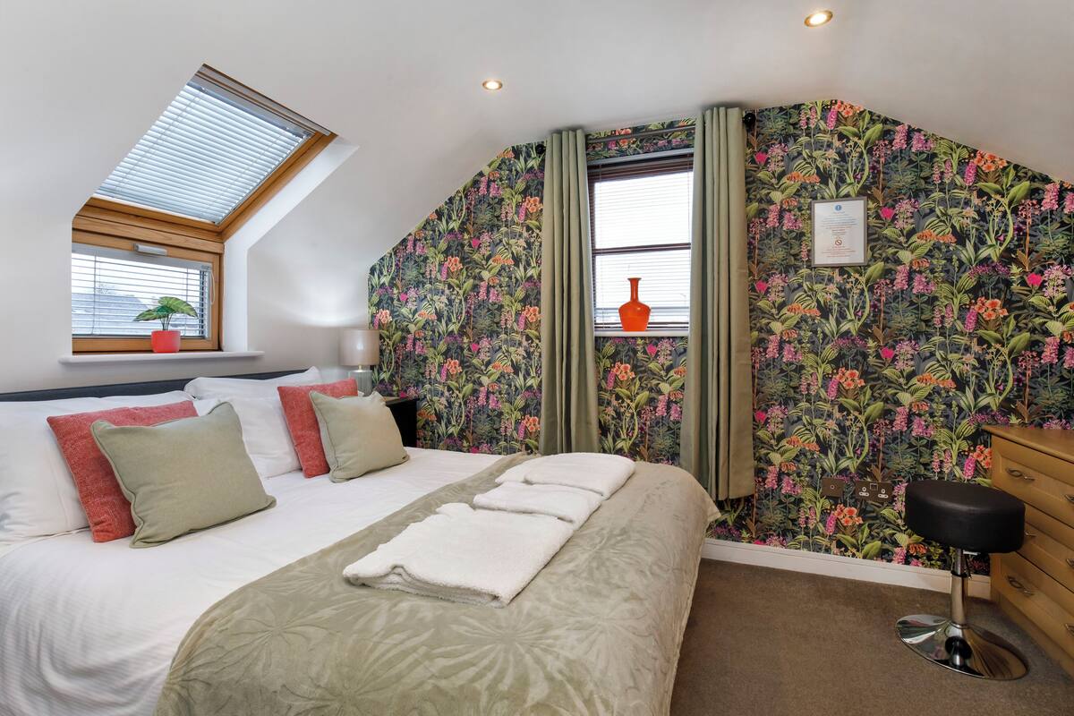 A bedroom with a skylight, floral wallpaper, a bed with multiple pillows, a bedside table, and a chair.