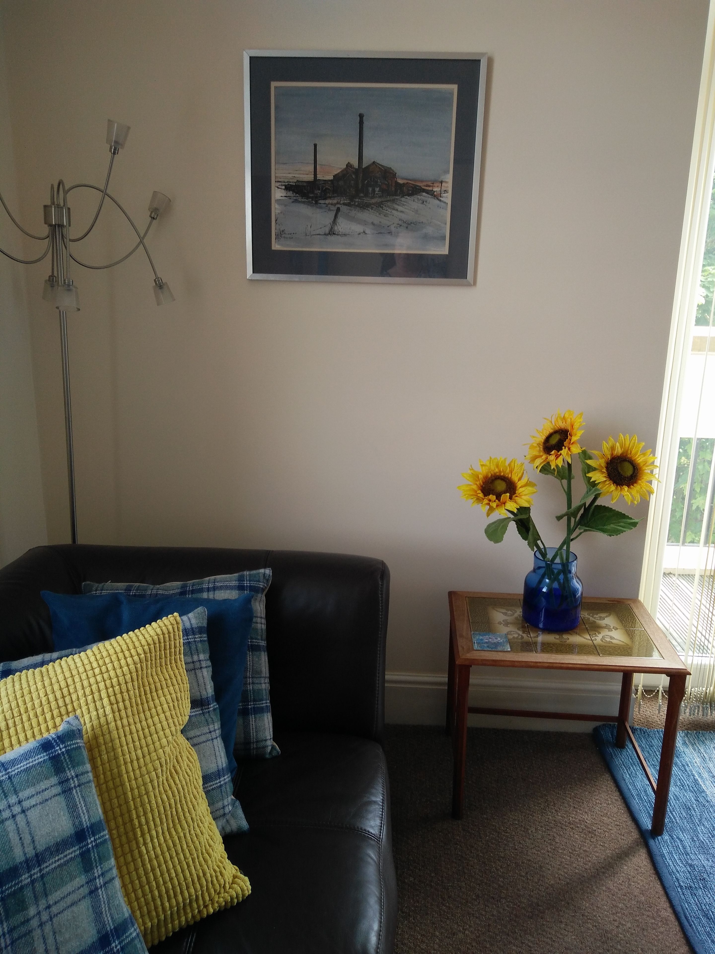 Un salón con un sofá de cuero negro, una mesita de madera con un jarrón de girasoles, y un cuadro en la pared.