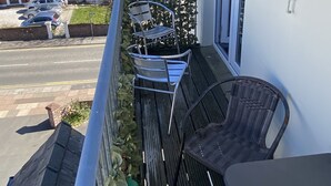 Un balcone con un tavolo e due sedie, vista su una strada con auto parcheggiate e un edificio con una torre dell'orologio sullo sfondo.