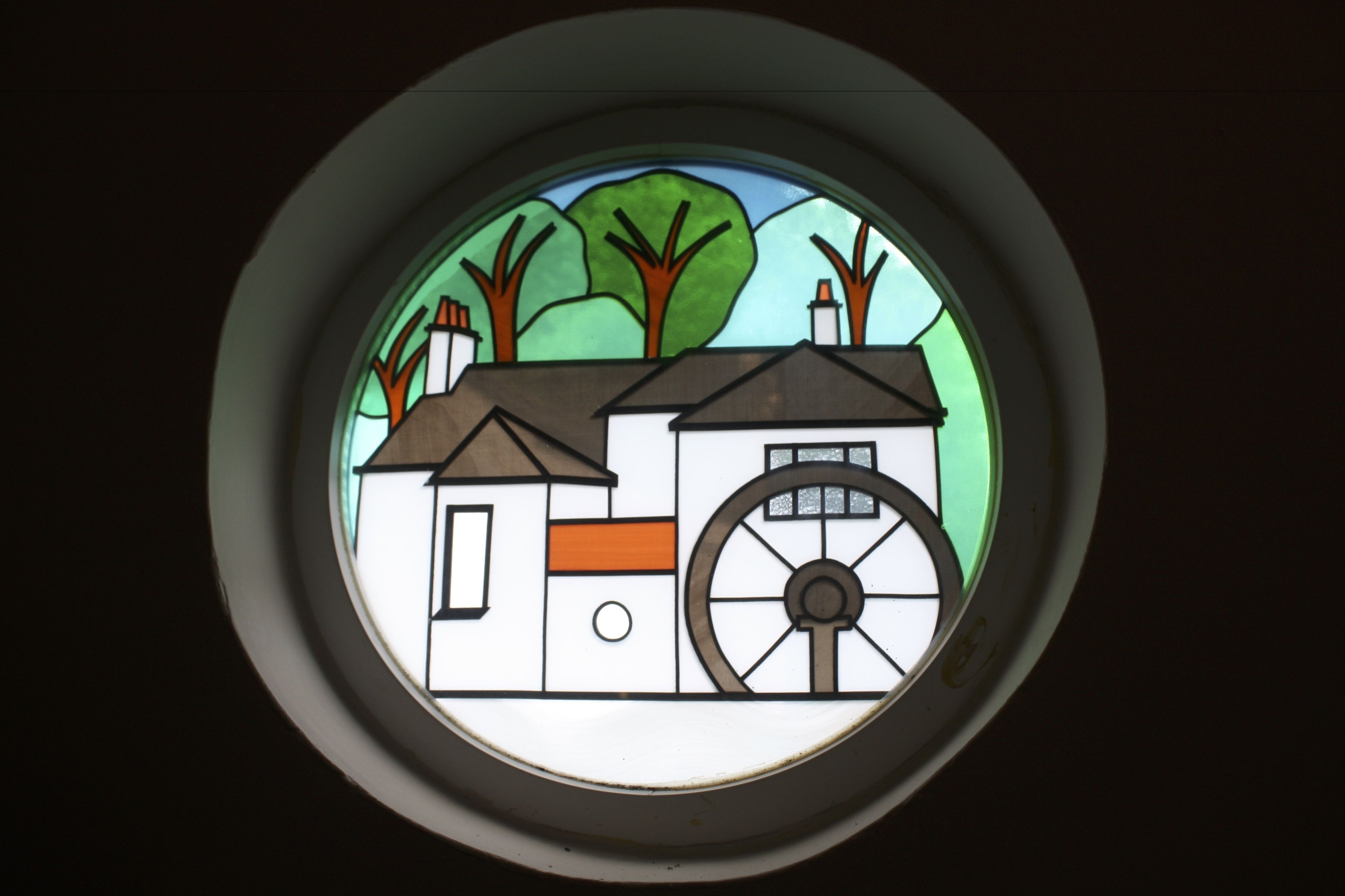 Uma janela redonda em vitral com uma casa e árvores.