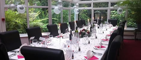 Una larga mesa dispuesta para una cena formal en un invernadero con paredes de vidrio, rodeada de sillas y velas.