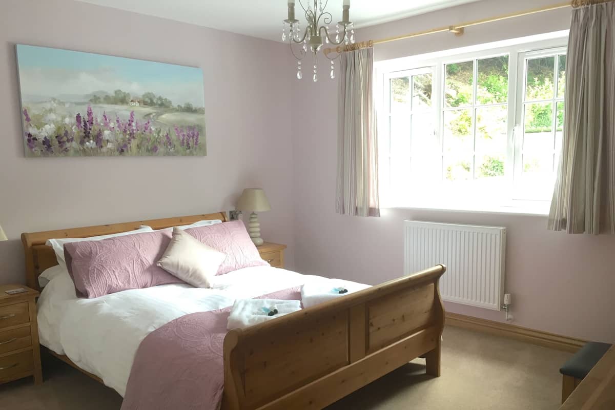 Ein Schlafzimmer mit einem hölzernen Bett, einem Nachttisch, einer Lampe, einem Fenster mit Vorhängen und einem Bild an der Wand.