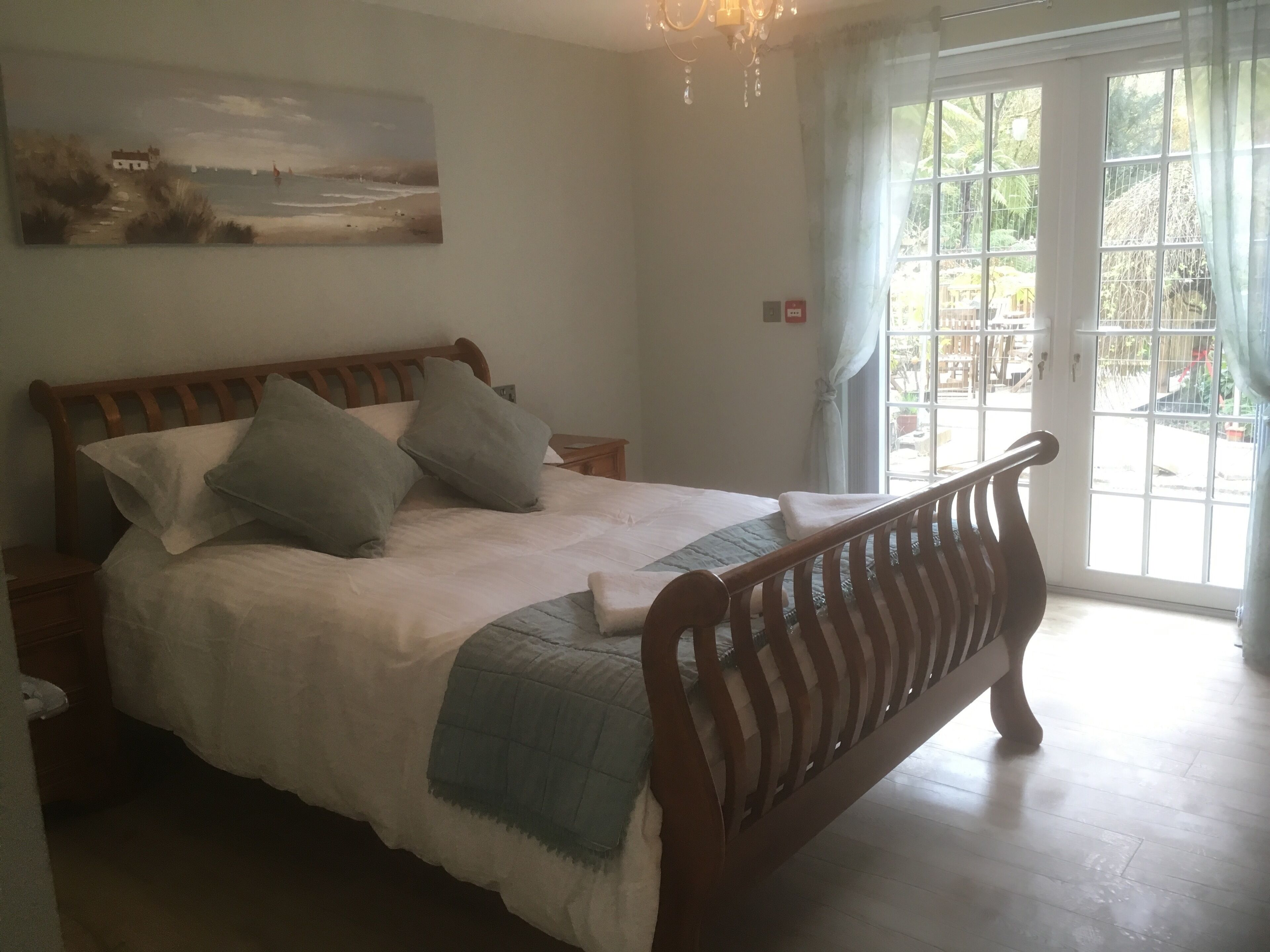 Une chambre avec un lit en bois, du linge de lit blanc, un plaid bleu, un tableau accroché au mur et une vue sur un jardin, visible à travers une grande fenêtre.