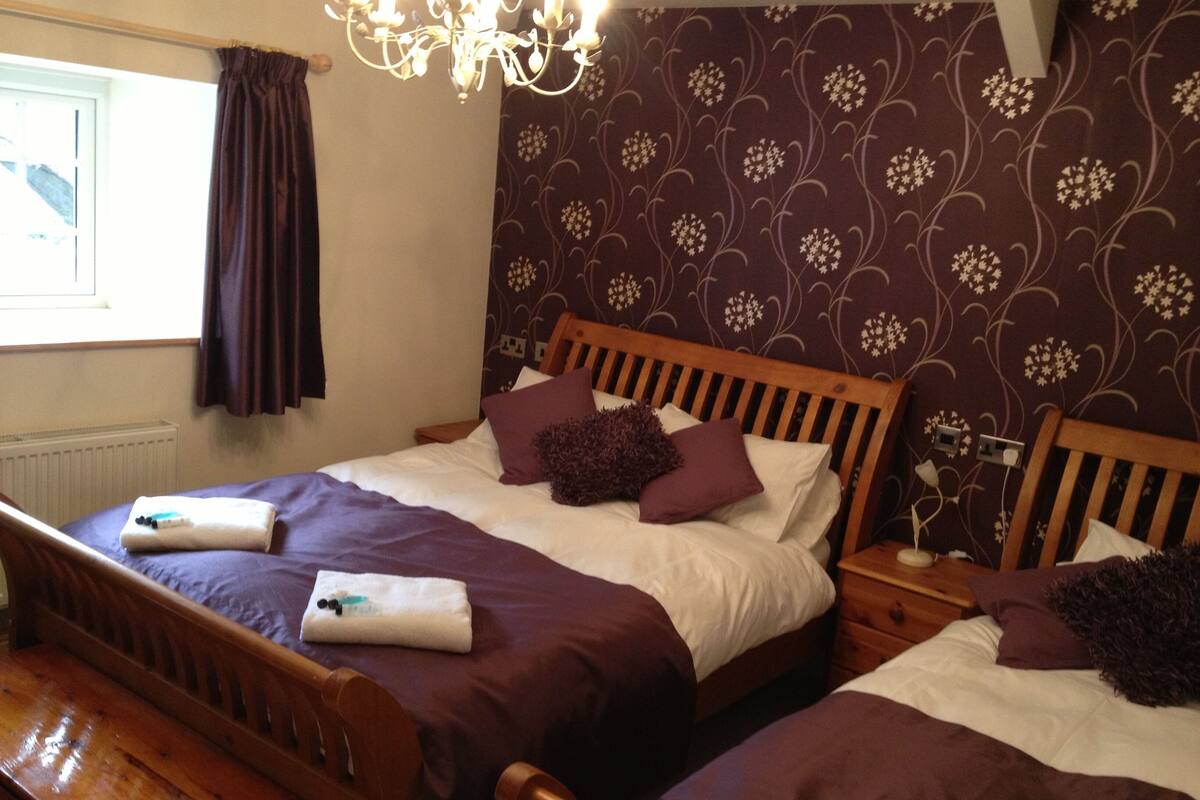 Ein Doppelbett mit Holz-Kopfteil, ein Nachttisch und ein Beistelltisch mit Lampe. Das Zimmer hat ein Fenster mit Vorhängen, eine Heizung und eine Wand mit Muster.