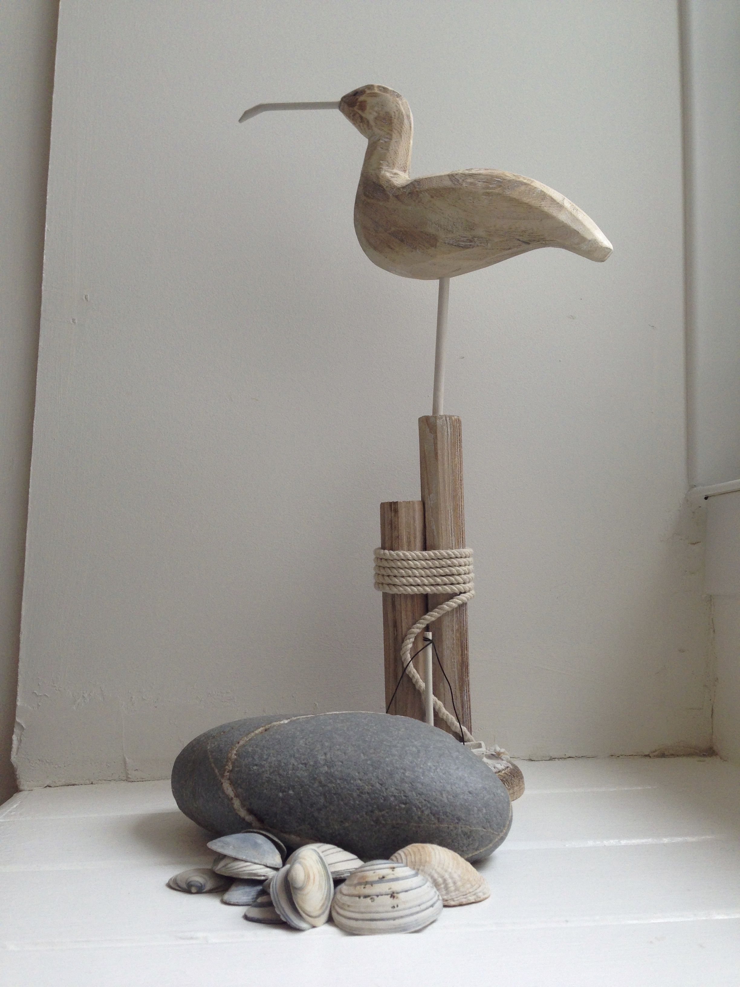 Eine Holzskulptur eines Vogels mit langem Schnabel, ein Seil, das um einen Holzpfosten gebunden ist, und mehrere Muscheln auf einer weißen Fläche.
