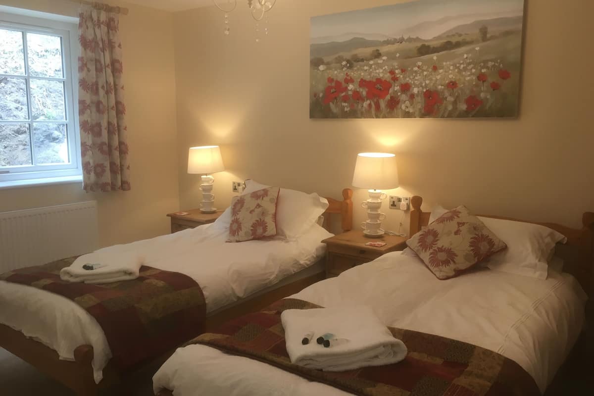 Ein Hotelzimmer mit zwei Einzelbetten, jeweils mit Nachttisch, Lampen und Kissen mit Blumenmuster. An der Wand hängt ein Gemälde eines Feldes mit roten Blumen.