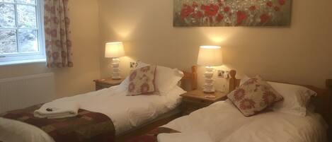 Habitación de hotel con dos camas individuales, cada una con mesita de noche, lámparas y almohadas estampadas con motivos florales. Un cuadro de un campo con flores rojas cuelga en la pared.