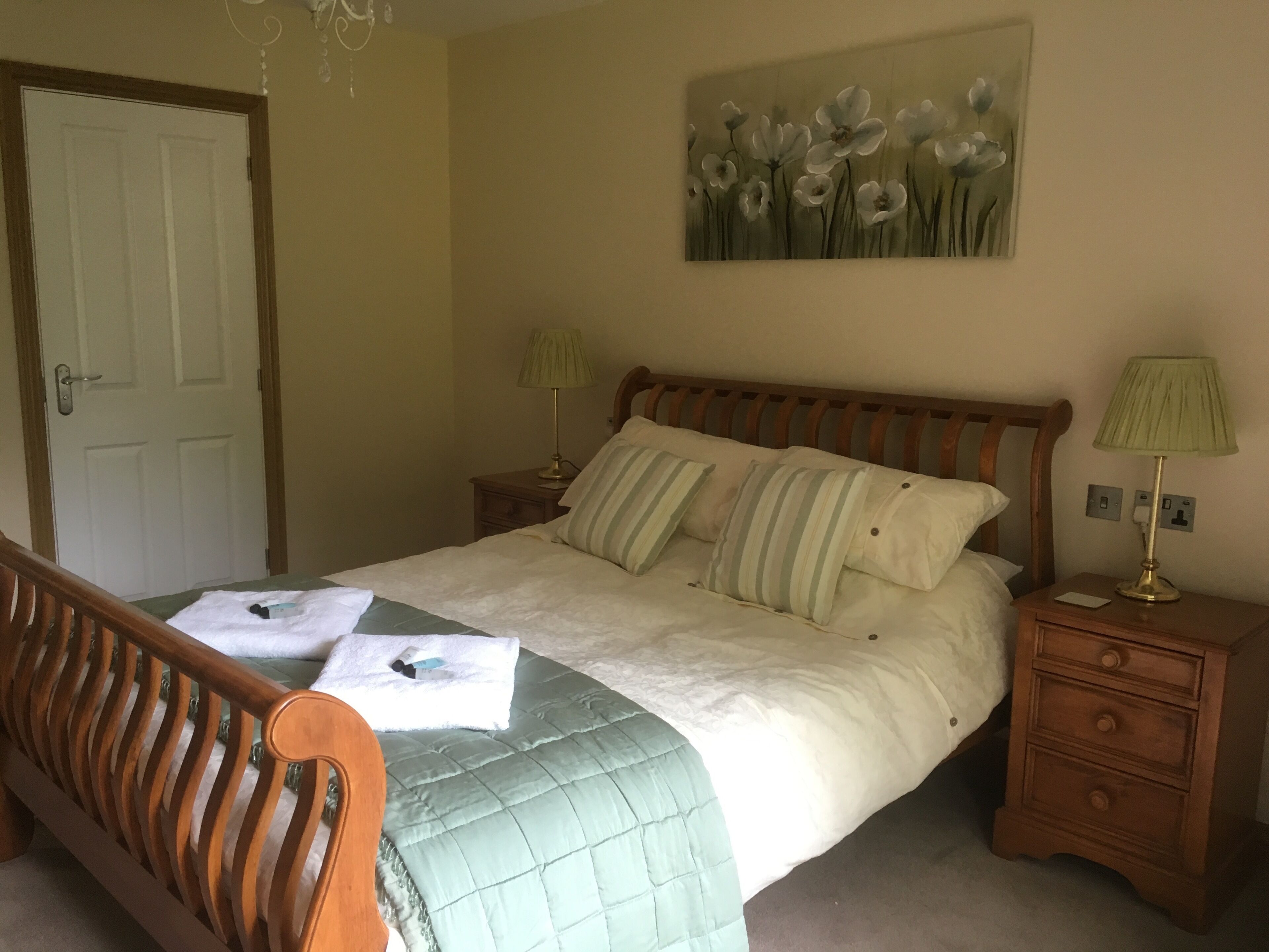 A neatly made bed with a green quilt, two bedside lamps, a wooden headboard, a painting of flowers on the wall, and a door in the background.