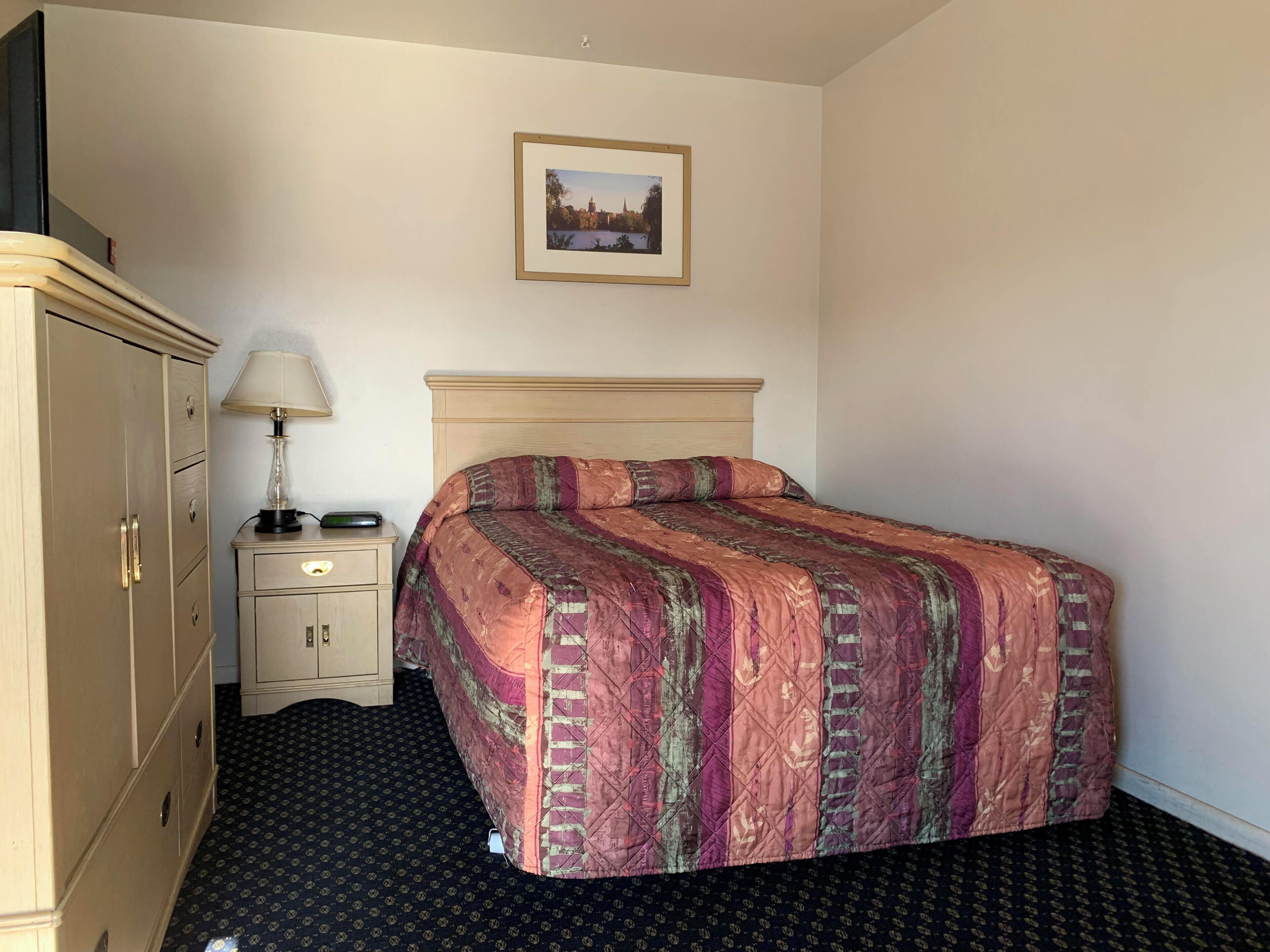 Camera d'albergo ordinata con un letto, un comodino con una lampada, un armadio e un quadro appeso al muro.