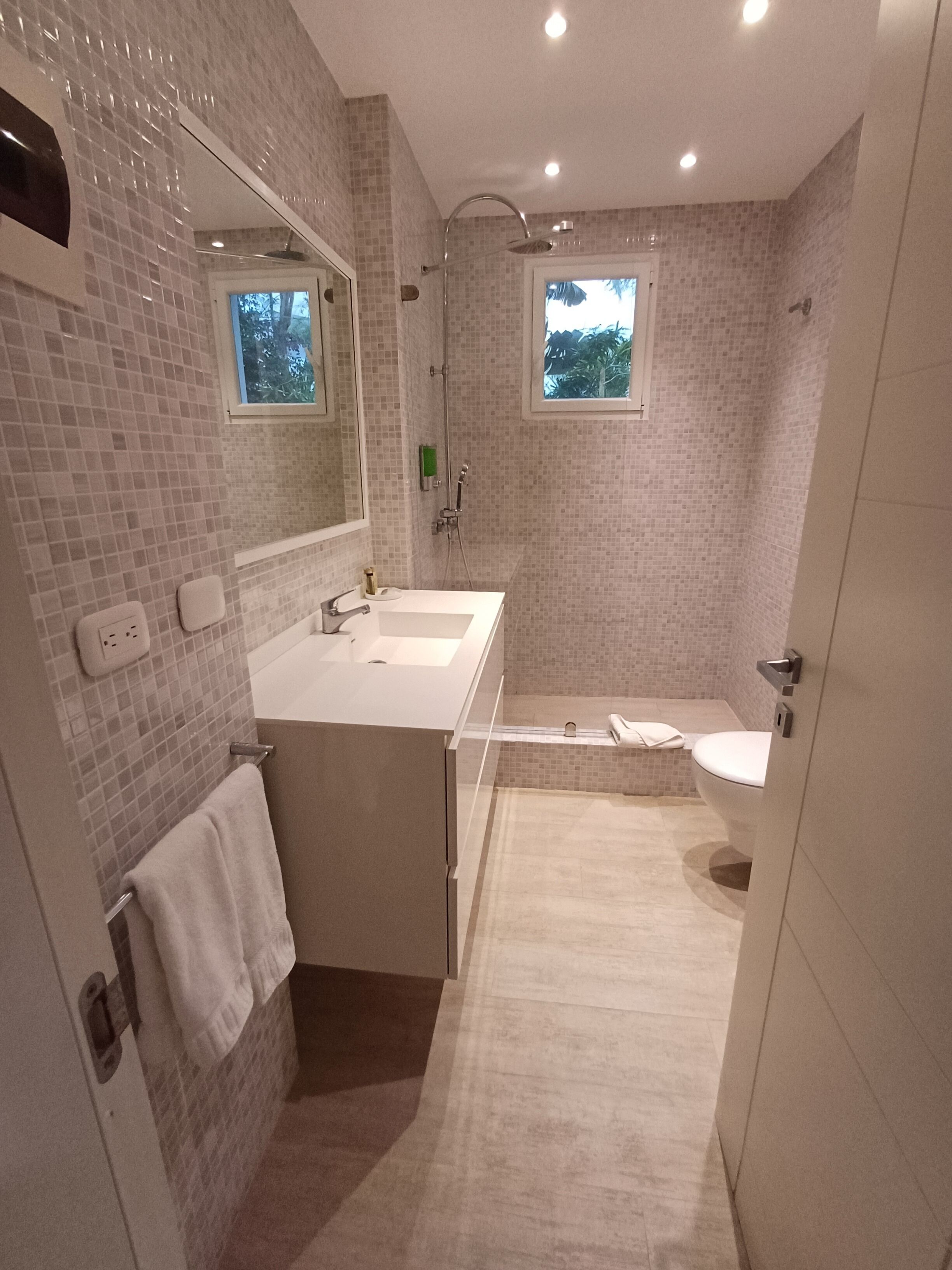 A modern bathroom with a large mirror, two sinks, a shower, and a toilet.