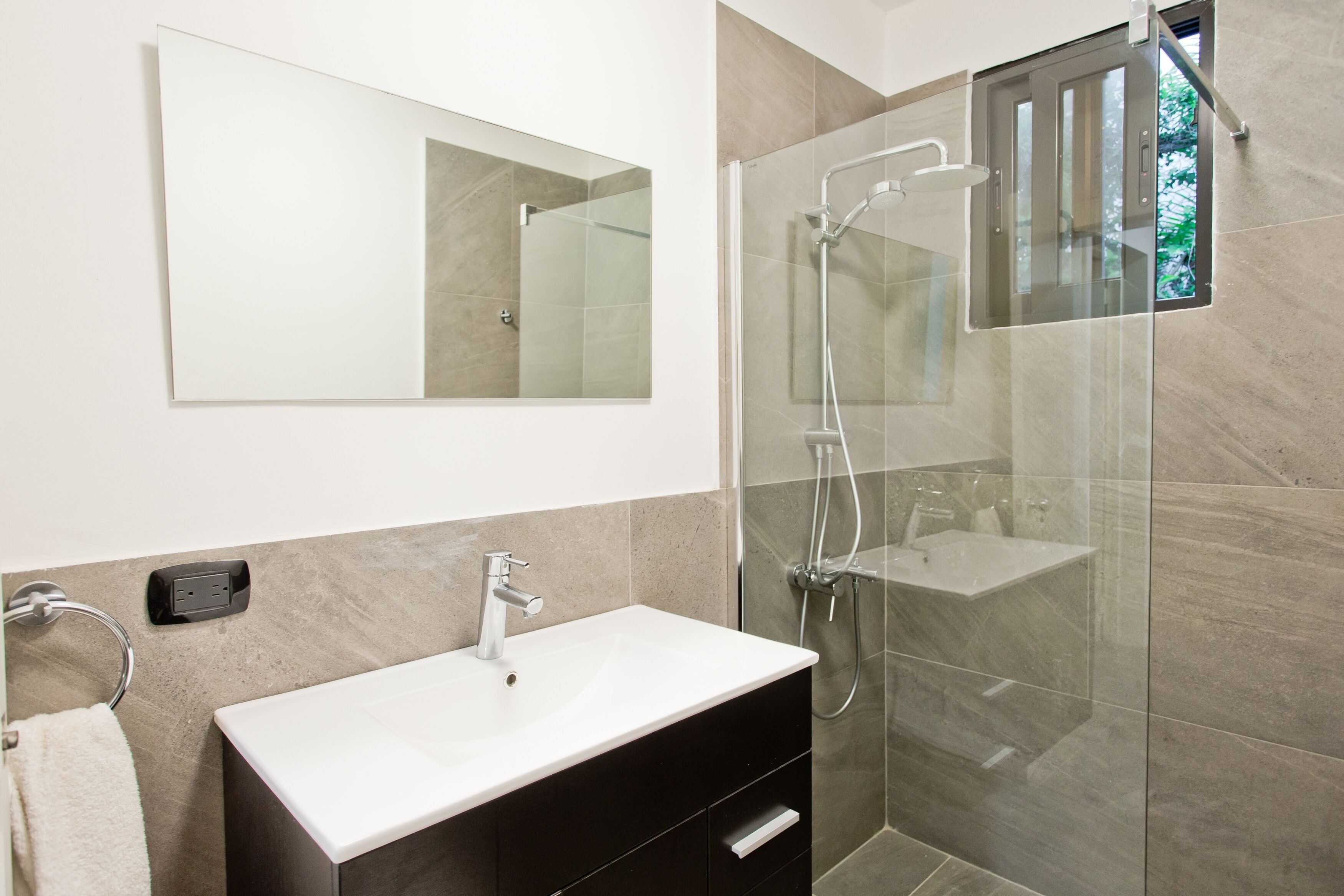 Un baño moderno con lavamanos blanco, mueble oscuro y ducha con cerramiento de vidrio.